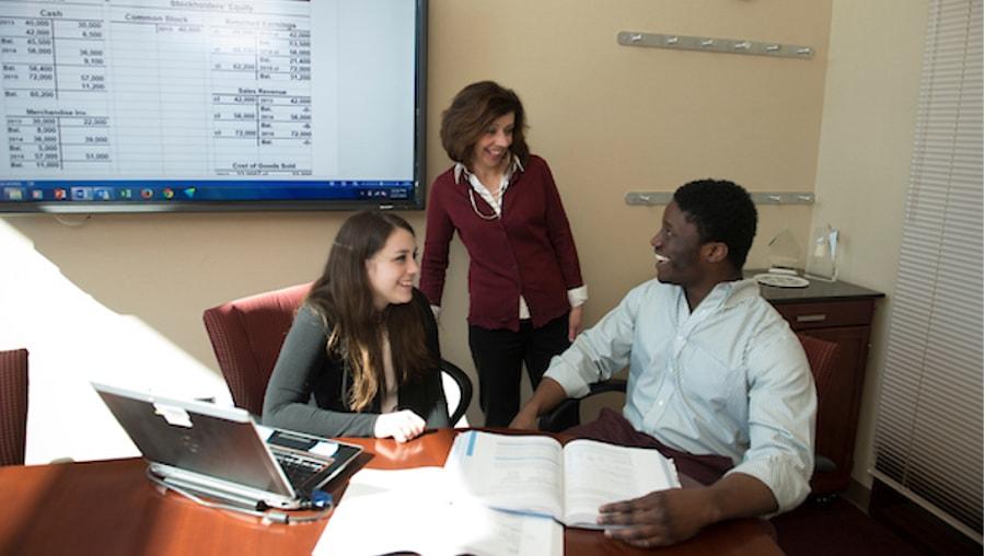 "Two students and an instructor conversing in a business setting classroom"