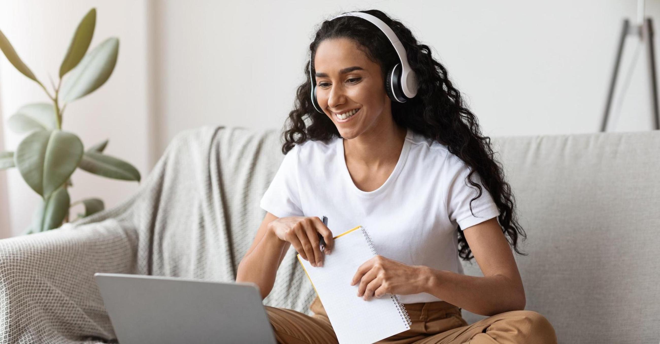 "A woman with headphones on, sitting at home on a couch, taking online classes"